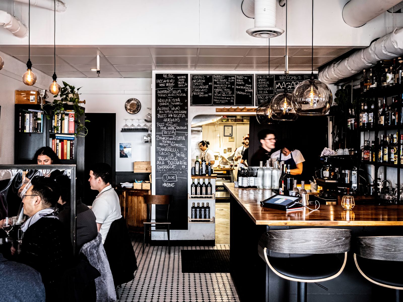 The dining room: chalkboard menus, the wooden bar with stools, penny-tile floor and the open kitchen behind